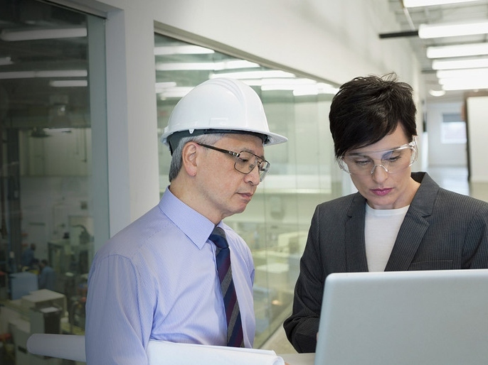 A man wearing a hard hat and glasses and a woman wearing glasses looking at a computer