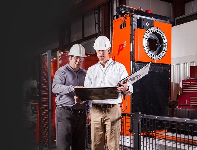 Two people in hard hats looking at a large binder with orange machine in background